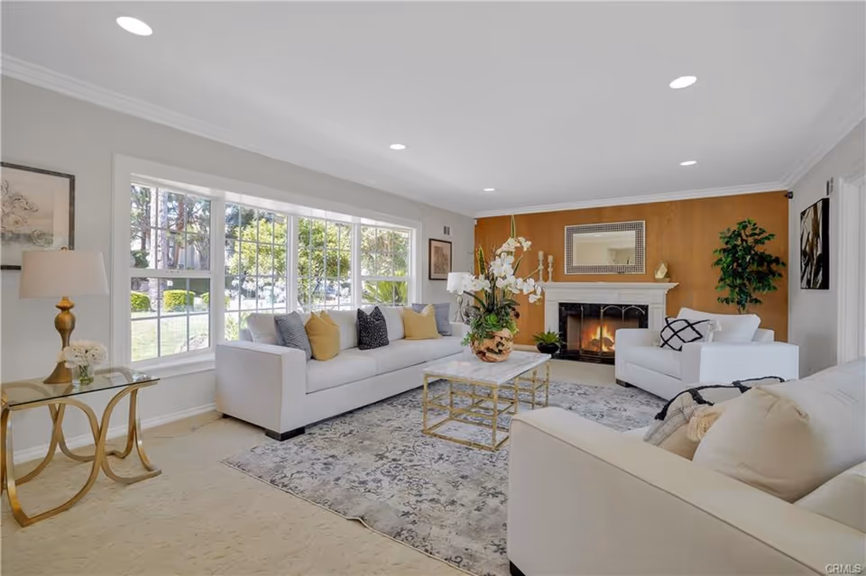 A bright and spacious living room with large windows letting in natural light. The room features white sofas with assorted cushions, a glass coffee table with a floral arrangement, a fireplace with a mirror above it, and a side table with a lamp and flowers. There is a patterned rug on the floor and a potted plant in the corner.