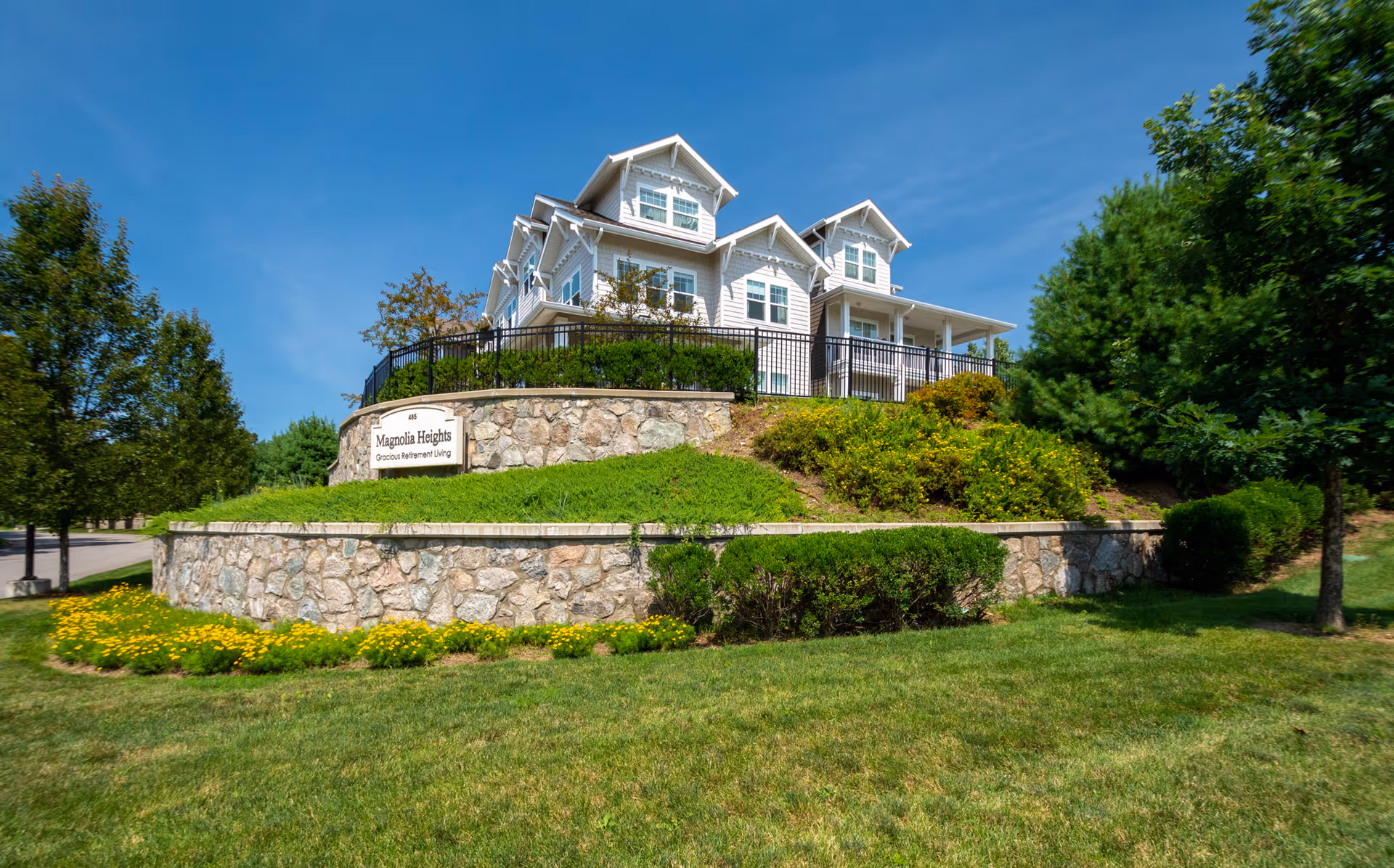 A white multi-story retirement building perched on a landscaped stone terrace with a sign reading "Magnolia Heights Gracious Retirement Living".