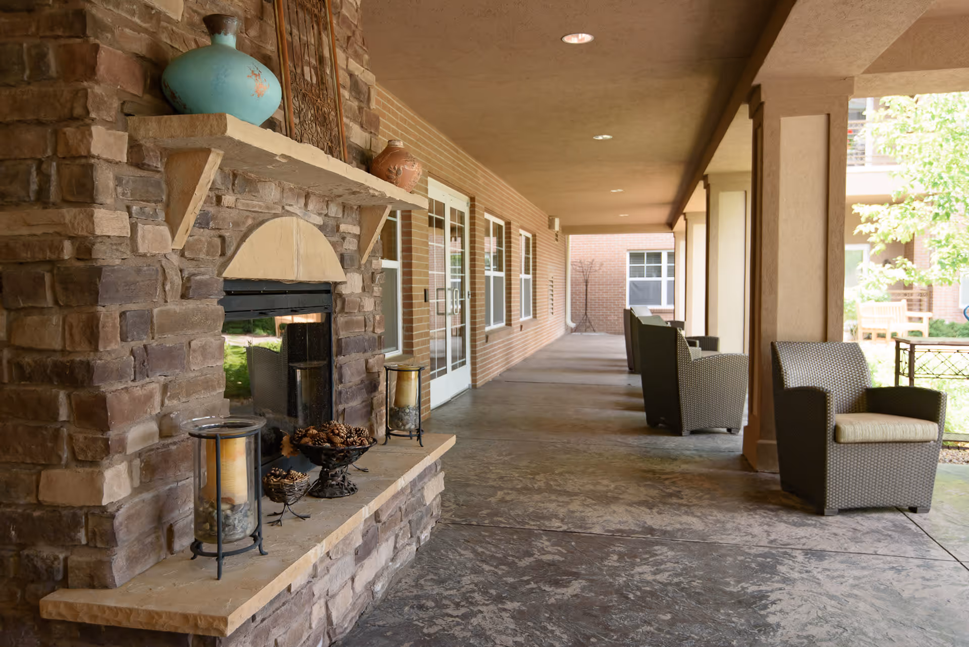 Covered outdoor patio area with stone fireplace decorated with vases and pine cones, wicker chairs with cushions arranged along the walkway, and brick walls with windows and glass doors leading inside.