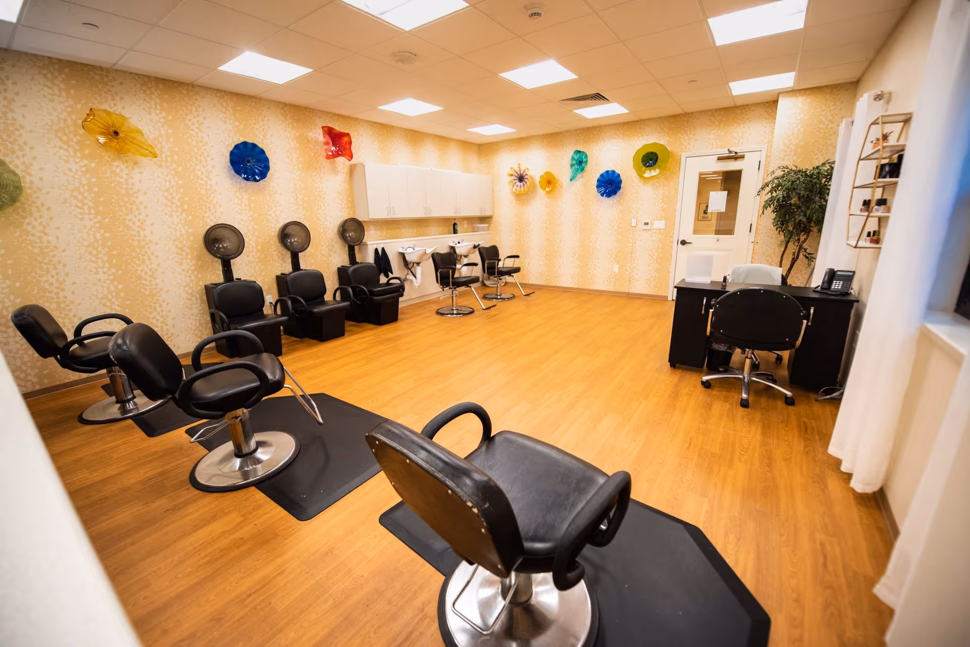 Interior view of a salon area in a senior living facility with multiple black salon chairs, hair drying stations, a desk with a chair, and colorful decorative wall art. The room has wood flooring and bright ceiling lights.