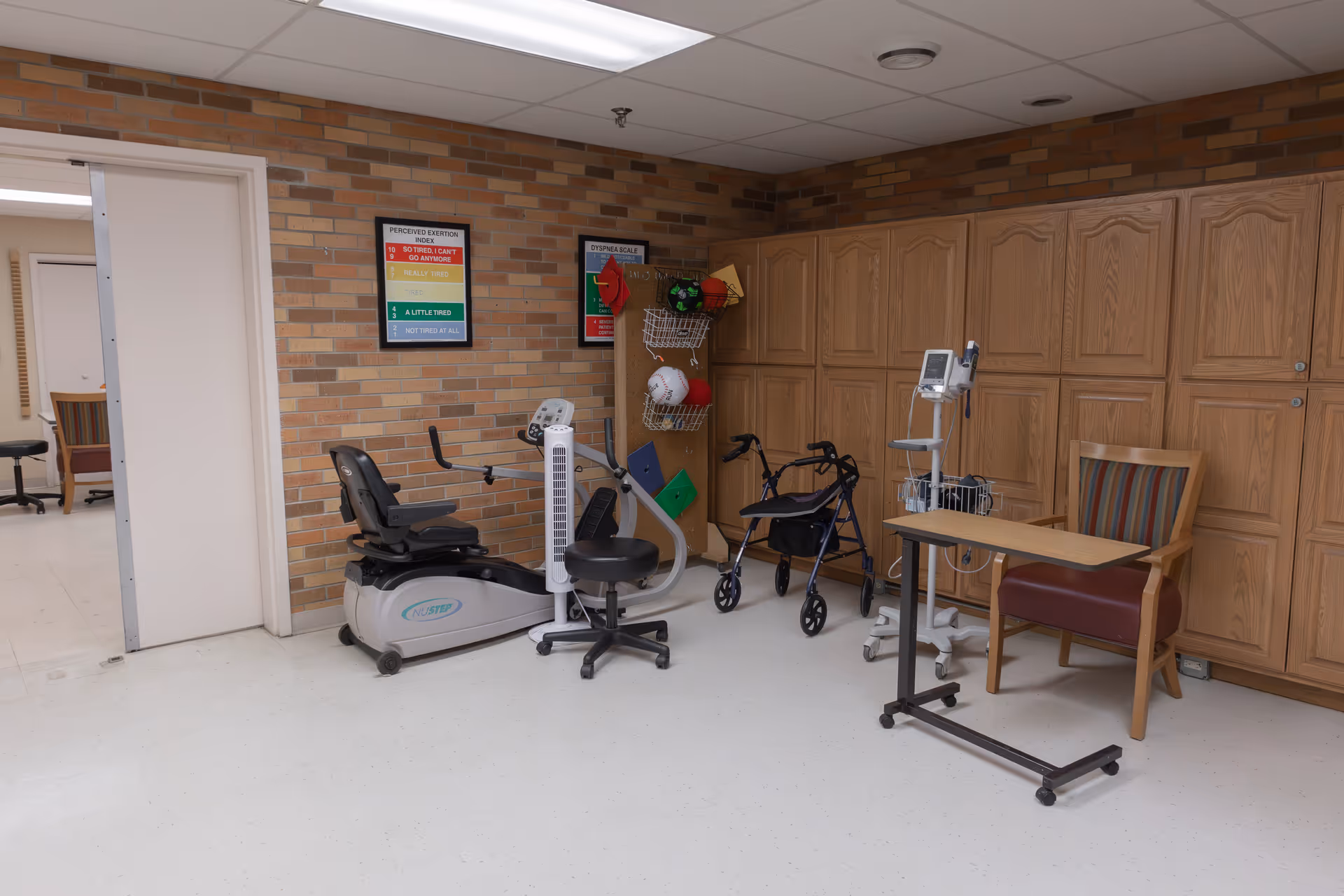 A room with exercise equipment including a recumbent bike, a walker, a medical monitor on a stand, a chair with a striped cushion, and a rolling table. The walls are brick with wooden cabinets along one side and two framed posters on the wall.
