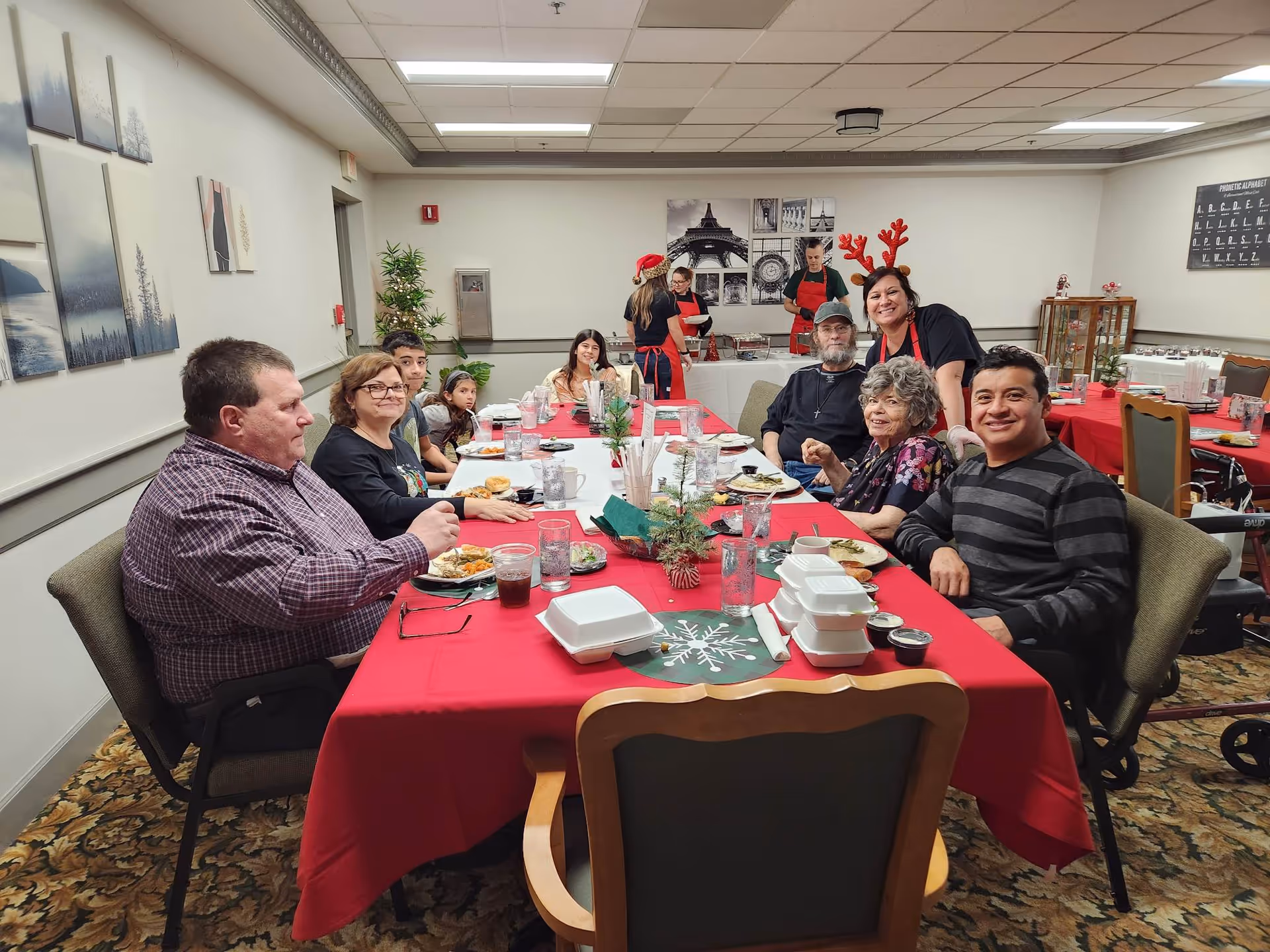 A group of people sit around a long red-covered table enjoying a meal in a decorated dining room.