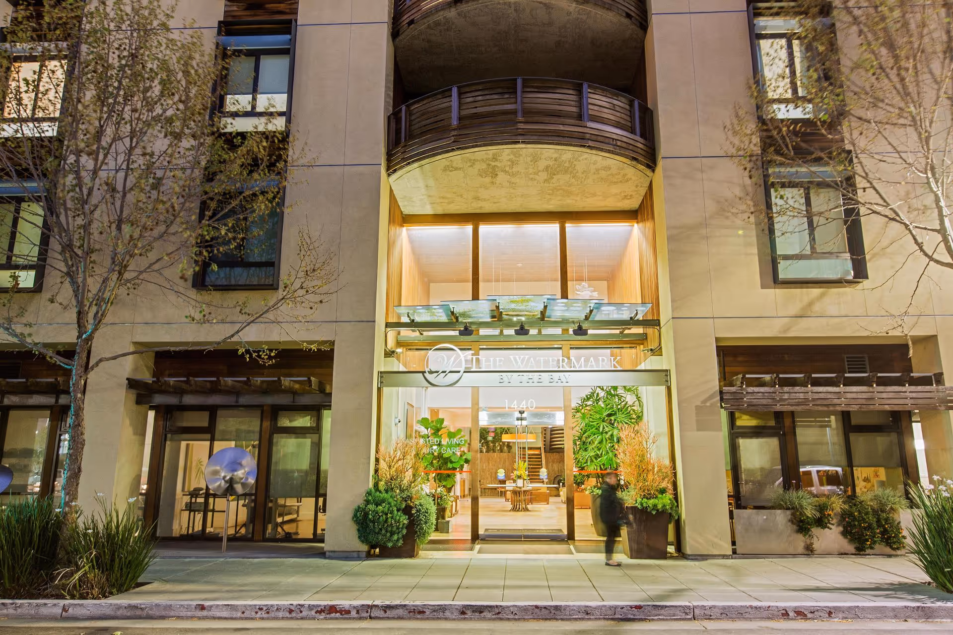 Illuminated front entrance of a senior living building (1440) with glass doors, potted plants, and a visible lobby at night.