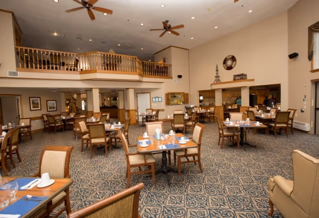 Spacious dining room with multiple wooden tables and chairs arranged neatly. Each table is set with placemats, cups, glasses, and utensils. The room has a high ceiling with ceiling fans and a balcony area overlooking the dining space. The walls are decorated with framed pictures and a large clock. There is a serving area or kitchen pass-through window on one side with people visible in the background.