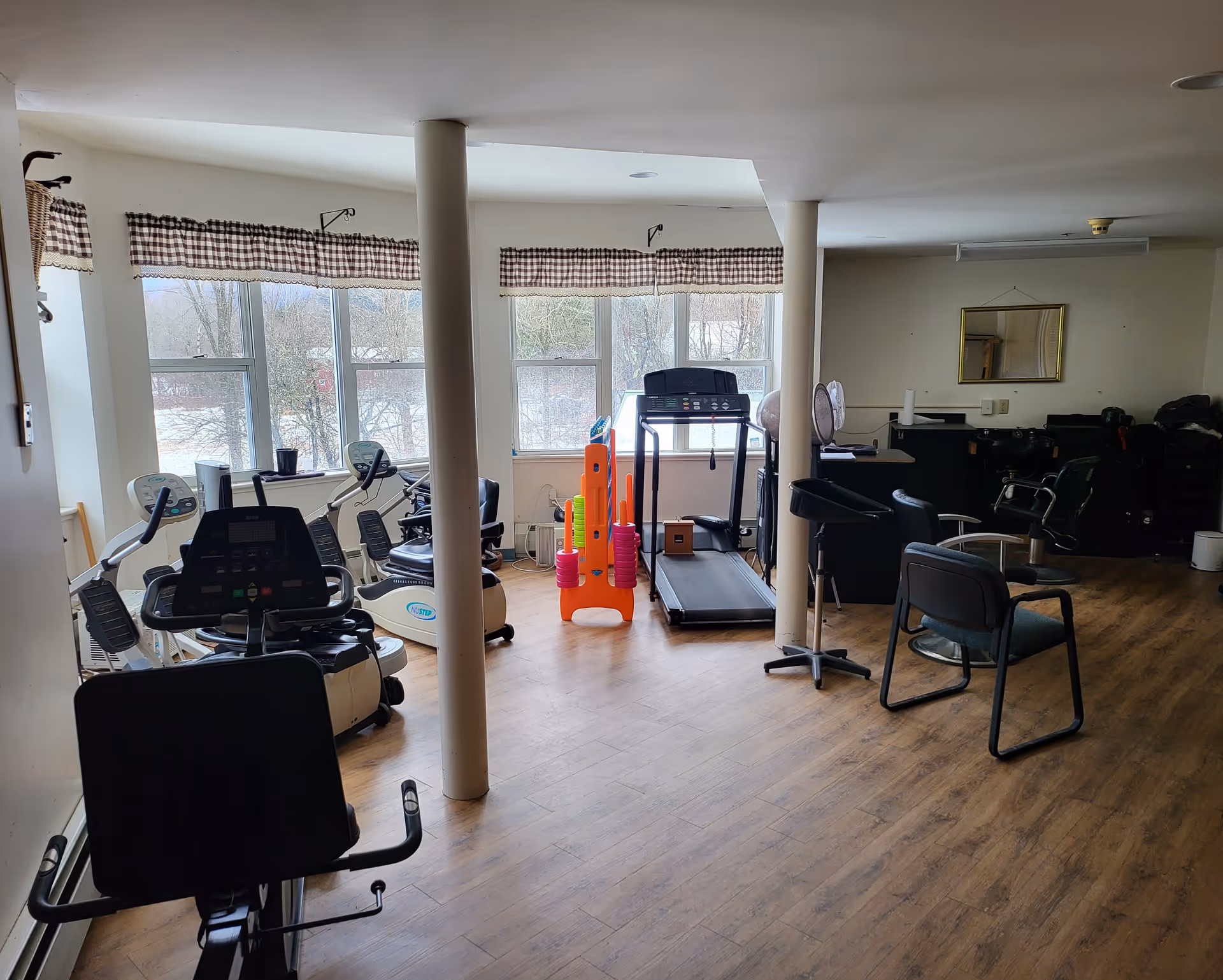 A room with exercise equipment including stationary bikes, a treadmill, and colorful exercise cones. The room has large windows with checkered valances, wooden flooring, and some chairs and fans. The space appears to be a fitness or activity room.