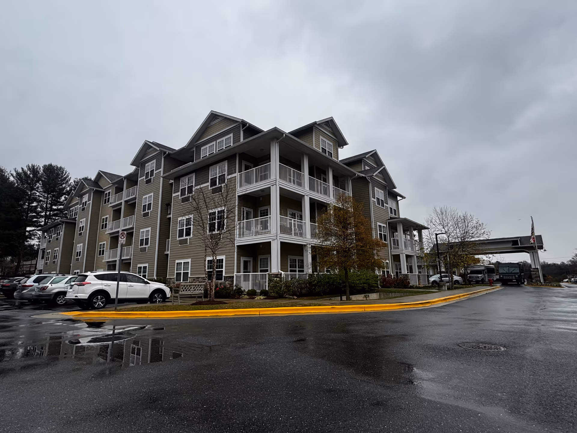 Exterior view of a multi-story residential building with balconies, parked cars, and a wet driveway under an overcast sky.