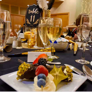 A close-up view of a dessert plate with chocolate-covered treats topped with pistachios, fresh berries, and a starfruit slice on a table set for dining. In the background, there are glasses of champagne, a table number sign reading 'table 11', and other dining table settings with people seated.