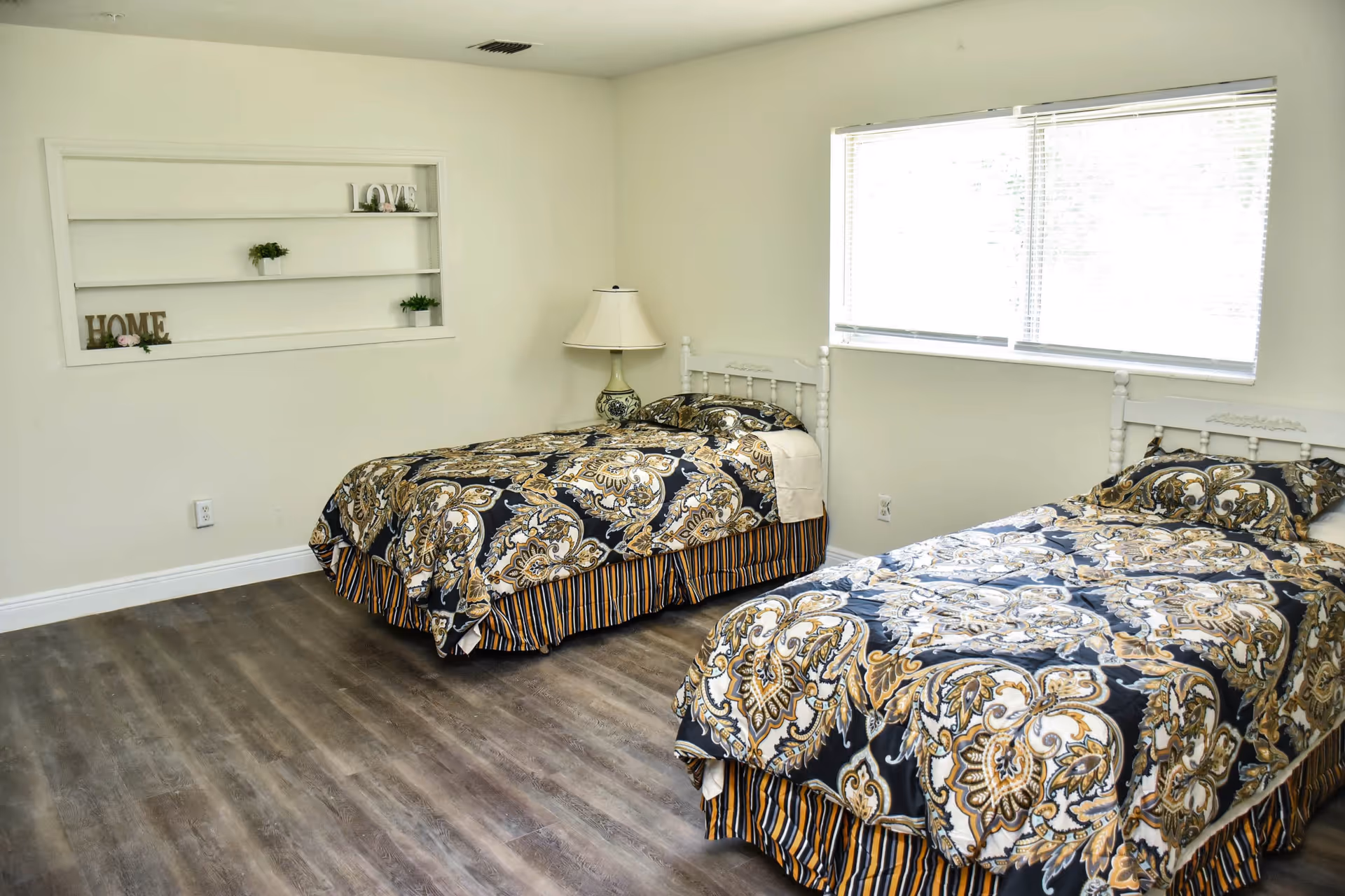 A bedroom with two twin beds featuring patterned bedspreads in black, gold, and white. There is a window with blinds on the right wall, a table lamp between the beds, and a built-in wall shelf with decorative items including the words 'LOVE' and 'HOME'. The floor is wood with a grayish tone.