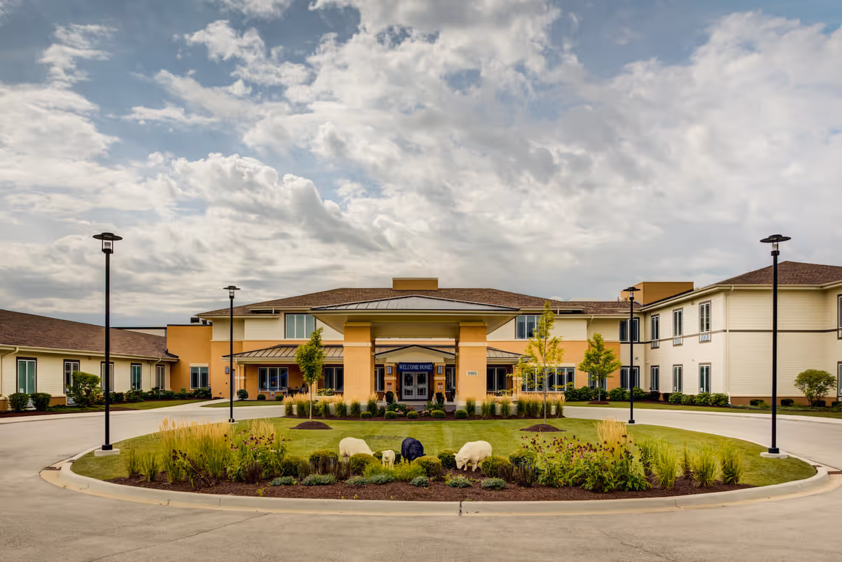 Front exterior view of Arbor Terrace Naperville, a two-story senior living facility with a covered entrance, surrounded by landscaped greenery and decorative sheep statues on a circular lawn under a partly cloudy sky.