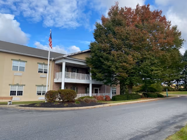 Exterior view of a two-story senior living facility building with beige walls and white trim. There is a covered entrance with white columns and a balcony above it. An American flag is flying on a flagpole in front of the building. A large tree with green and reddish leaves is near the entrance, and there is a curved driveway in front of the building.