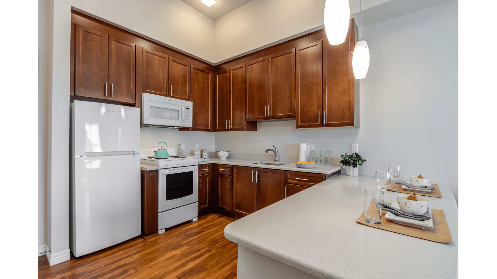 Modern kitchen with dark wood cabinets, white appliances including a refrigerator, stove, and microwave, and a light-colored countertop with two place settings. The floor is wooden, and two pendant lights hang above the counter.