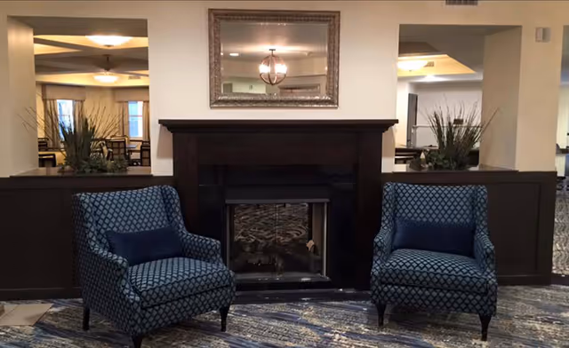 A cozy seating area with two patterned blue armchairs facing a dark wooden fireplace. Above the fireplace is a large framed mirror reflecting a chandelier and parts of the room. The space features neutral walls, decorative plants on either side of the fireplace, and a patterned carpet.