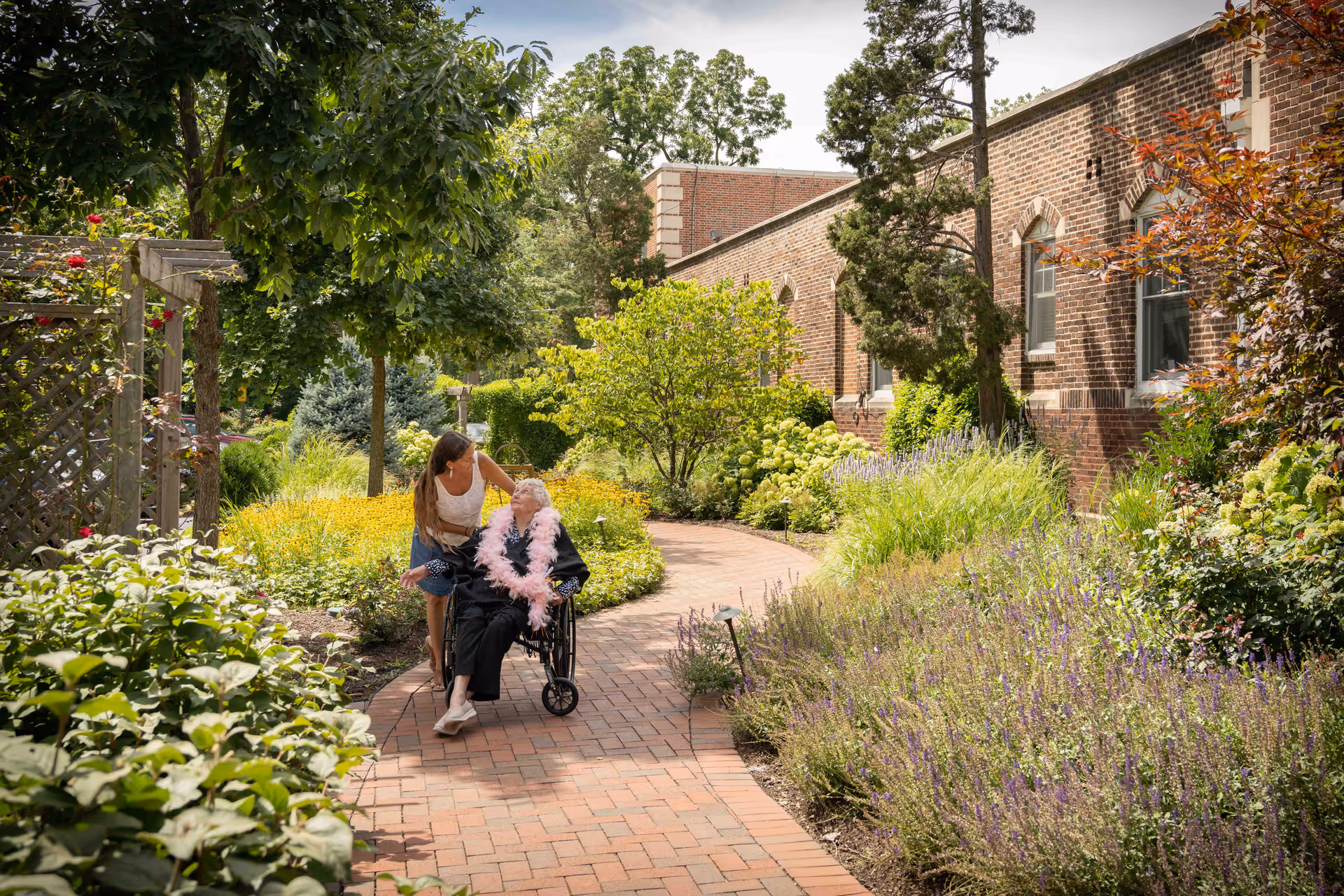 A woman pushing an elderly person in a wheelchair along a brick pathway in a lush garden area next to a brick building. The elderly person is wearing a pink feather boa and both appear to be enjoying the outdoor setting surrounded by greenery and flowers.