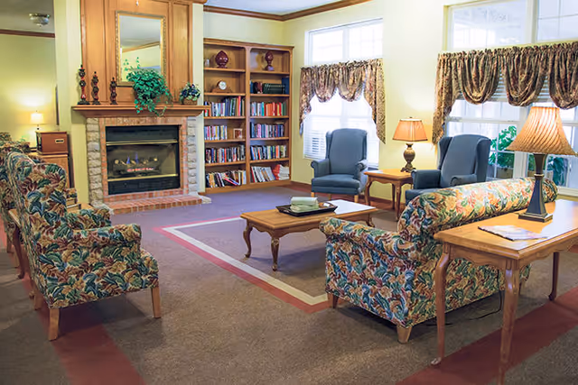 A cozy living room with floral patterned armchairs and a matching sofa arranged around a wooden coffee table on a carpeted floor. There is a fireplace with a wooden mantle and stone surround, flanked by built-in bookshelves filled with books and decorative items. Two blue armchairs are positioned near windows with patterned valances, and two table lamps provide warm lighting.