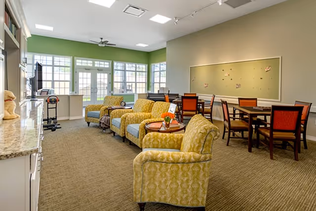 A bright and spacious common area in a senior living facility with several yellow patterned armchairs arranged around small round tables, a dining table with chairs in the background, large windows letting in natural light, and a bulletin board on the wall.