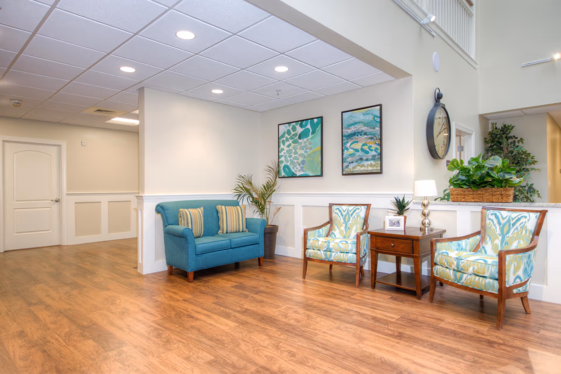 A bright and clean senior living facility common area with wooden flooring, a teal loveseat with two striped cushions, two patterned armchairs, a wooden side table with a lamp and small plant, two abstract paintings on the wall, a large wall clock, and several potted plants.