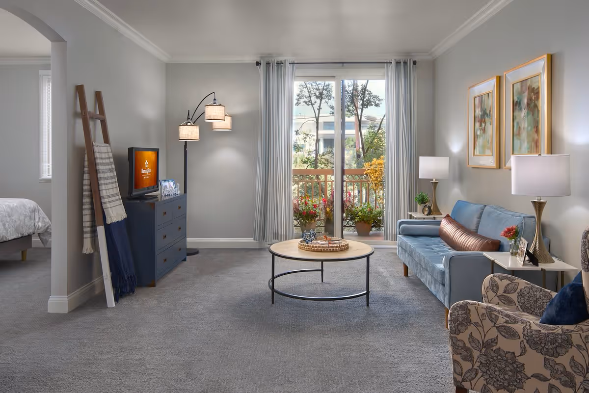 A cozy living room with a blue sofa, a round wooden coffee table, a patterned armchair, and two framed paintings on the wall. There is a floor lamp with three shades, a TV on a blue dresser, and a ladder-style blanket rack. Large sliding glass doors with curtains open to a balcony with potted plants and trees visible outside. A bedroom is partially visible through an arched doorway on the left.