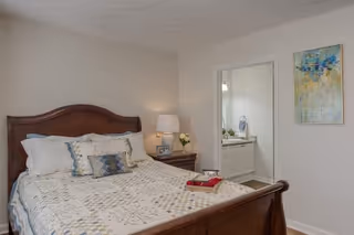 A bedroom with a wooden bed frame, a quilted bedspread, and decorative pillows. There is a nightstand with a lamp and a small flower vase. A doorway leads to a bathroom with a sink and mirror. A colorful abstract painting hangs on the wall.