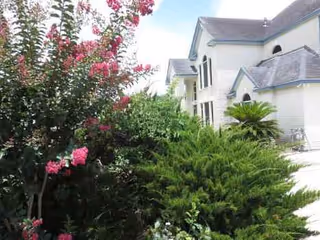White multi-story senior living building with peaked roofs and lush flowering shrubs in the foreground.