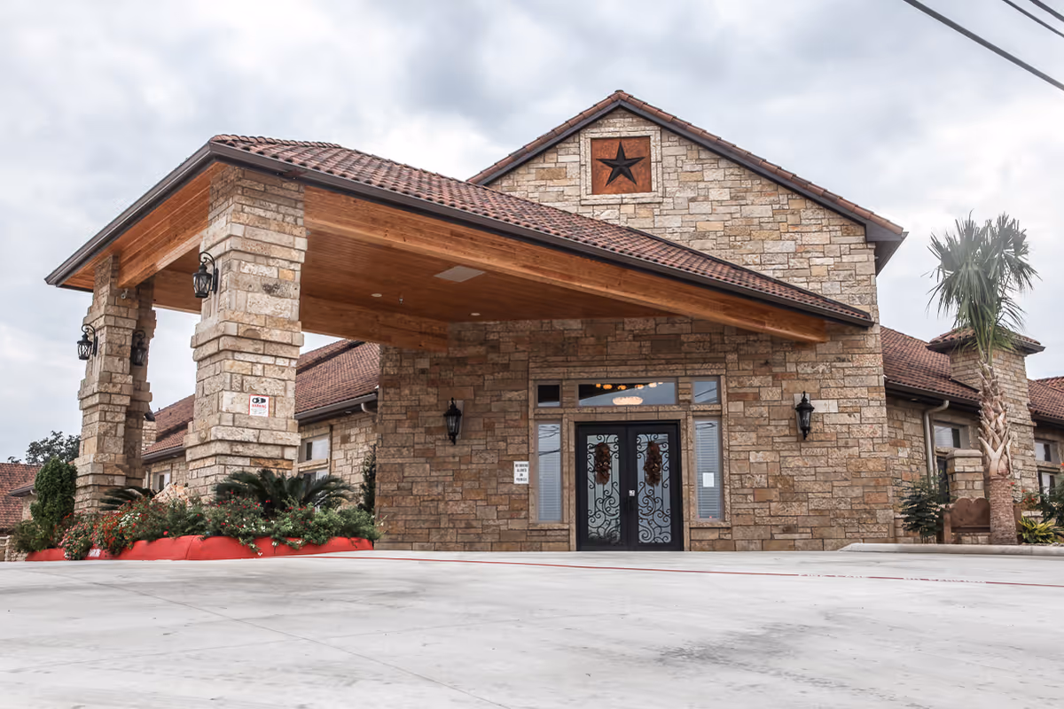 Front exterior view of Heritage Creek Assisted Living facility showing a stone building with a covered entrance supported by stone pillars, decorative black wrought iron double doors, and a star emblem above the entrance. There are plants and palm trees around the building and a cloudy sky in the background.