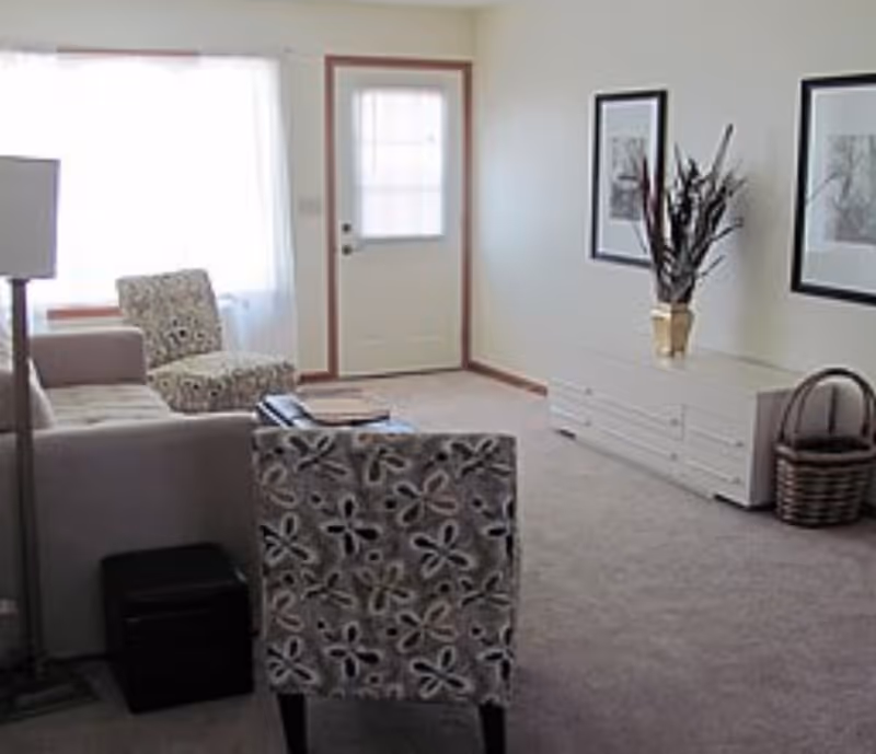 A bright living room with a sofa and patterned chairs, a floor lamp, a front door, and a low dresser topped with a plant and framed artwork.