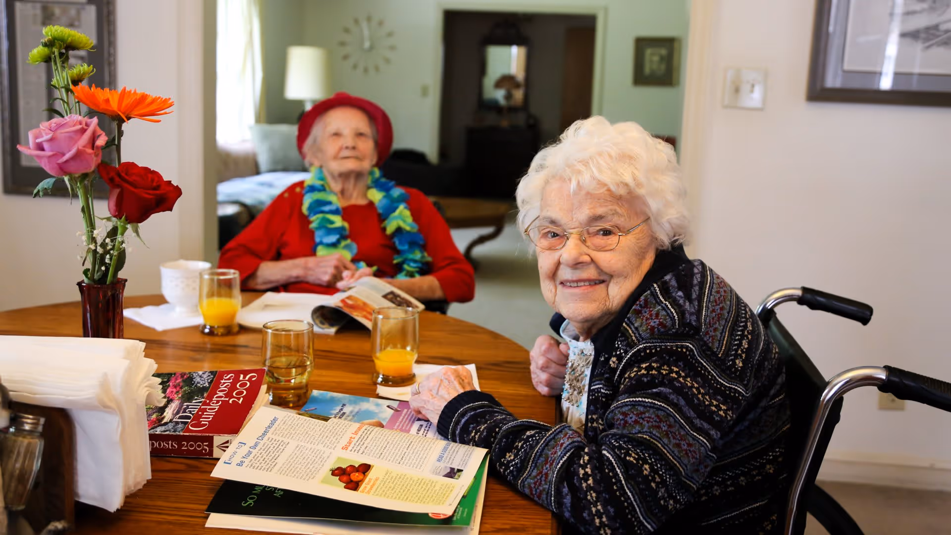 Two elderly women seated at a dining table with flowers, magazines, and glasses of orange juice, one smiling toward the camera.