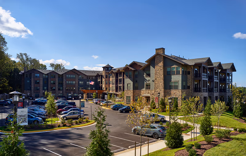 Exterior view of a multi-story senior living facility with stone and siding facade, a parking lot, and landscaped grounds under a blue sky.