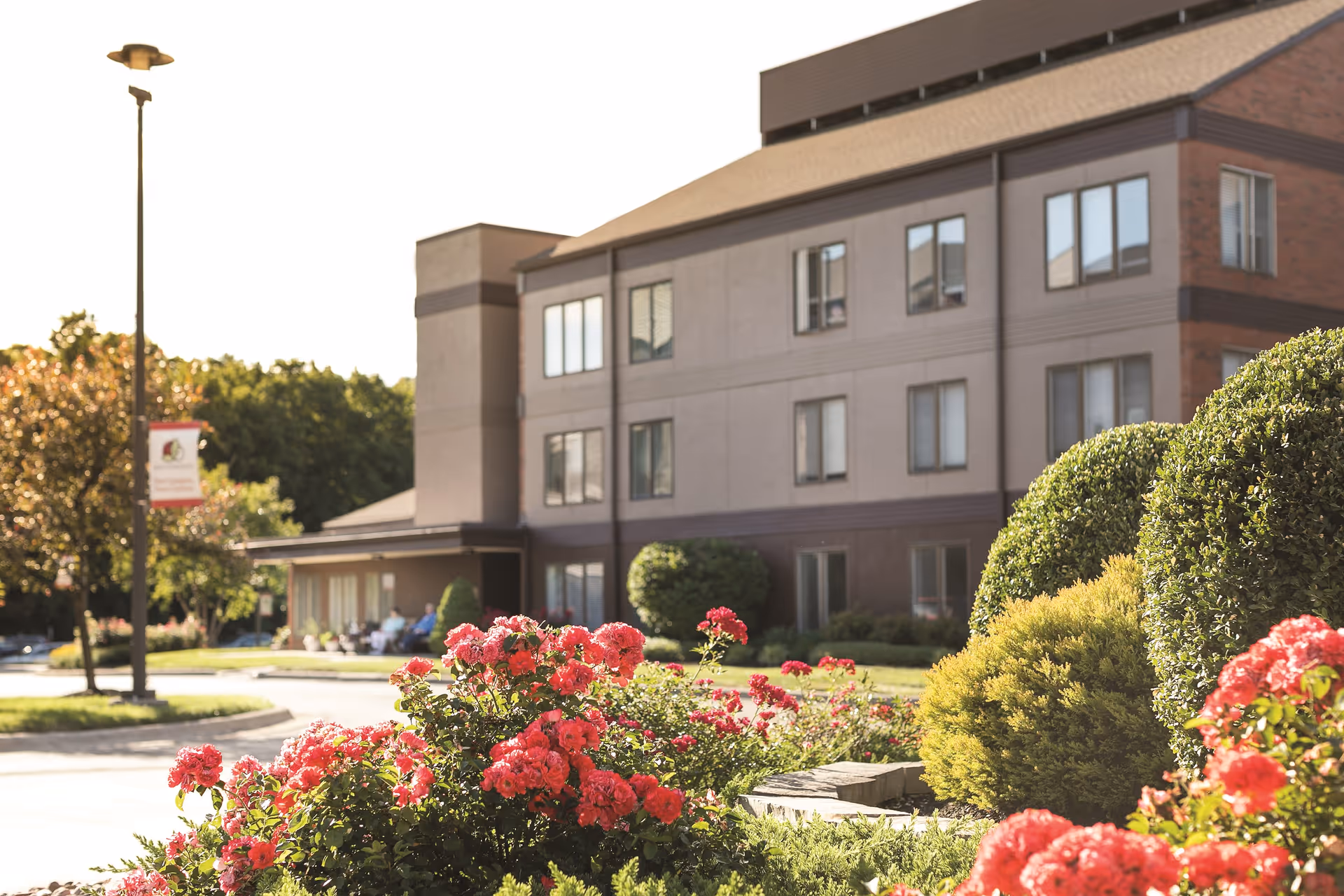 Exterior view of a multi-story senior living facility building named Westerwood with well-maintained landscaping including bushes and blooming red flowers in the foreground, and a street lamp with a banner on the left side.