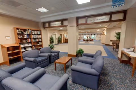 A spacious indoor common area with blue upholstered armchairs arranged around a wooden coffee table. There are bookshelves filled with books on the left side and several potted plants placed around the room. The area has carpeted flooring and a ceiling with recessed lighting. Signs above the columns indicate directions to different rooms.