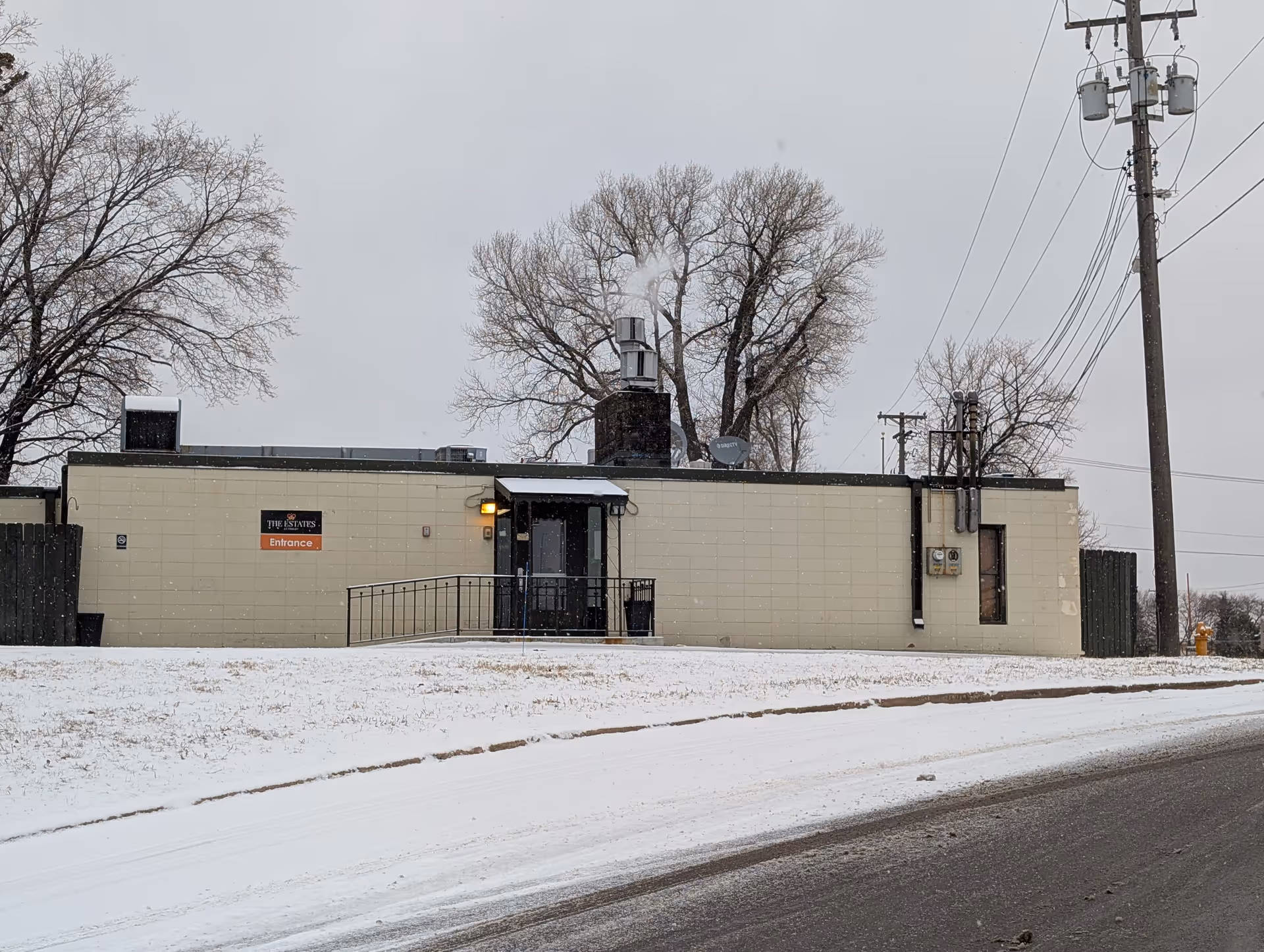 Exterior view of a single-story beige building with a black door entrance and a small ramp leading up to it. The building has a sign that reads 'The Estates Entrance'. There is snow on the ground and leafless trees in the background under a gray sky.