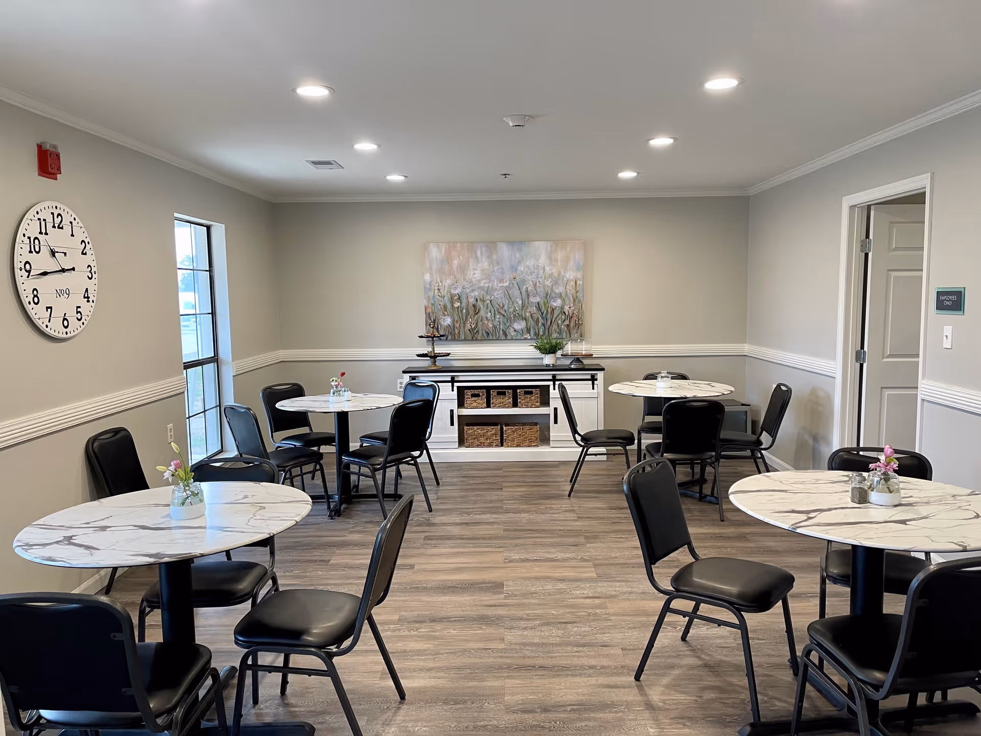 A small dining room with four round tables, each surrounded by black chairs. The tables have white marble-patterned tops and small flower vases as centerpieces. The room has light gray walls with white trim, a large wall clock, a window, and a sideboard with baskets and a decorative painting above it. The floor is wood-style laminate.