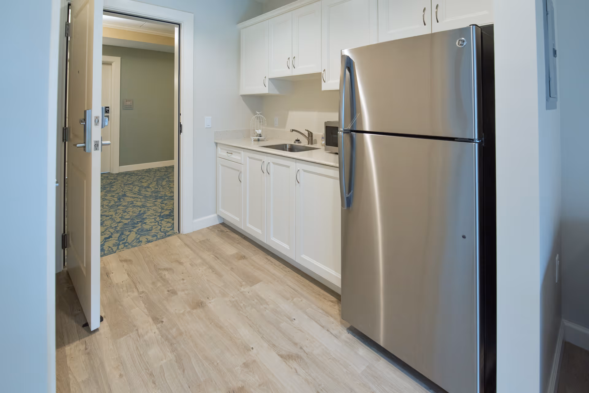 A small kitchen area with white cabinets, a stainless steel refrigerator, a sink, and a microwave. The floor is light wood, and an open door leads to a carpeted hallway with a blue and beige patterned carpet.