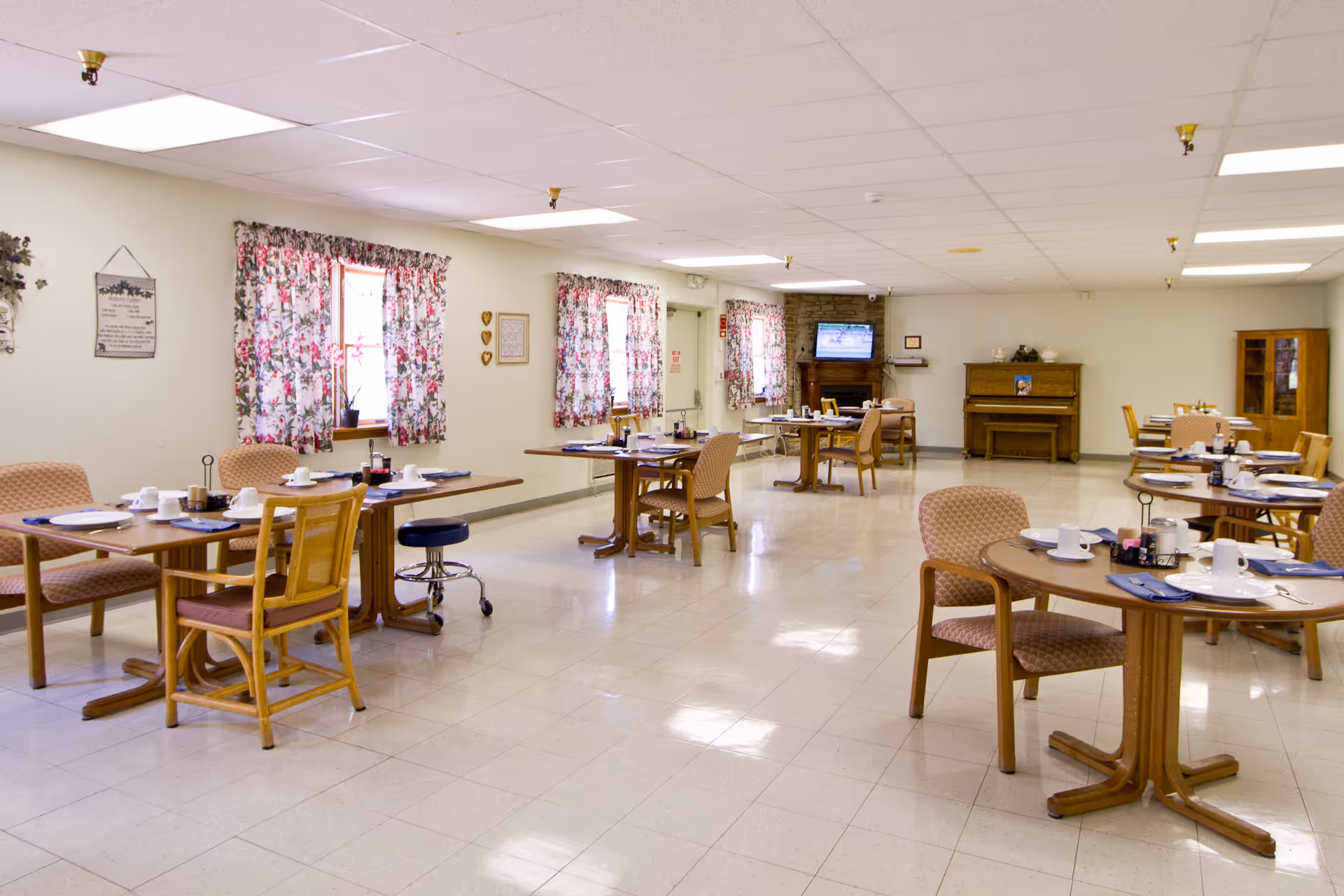 A bright dining room in a senior living facility with multiple wooden tables and chairs arranged neatly. Each table is set with white cups, plates, and napkins. The room has floral curtains on the windows, a piano against the far wall, and a television mounted above a fireplace. The floor is tiled, and the ceiling has recessed lighting.