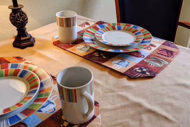 A dining table set for two with colorful striped plates and matching mugs on patterned placemats. A decorative lamp is visible on the left side of the table, and a dark chair is positioned at the table.