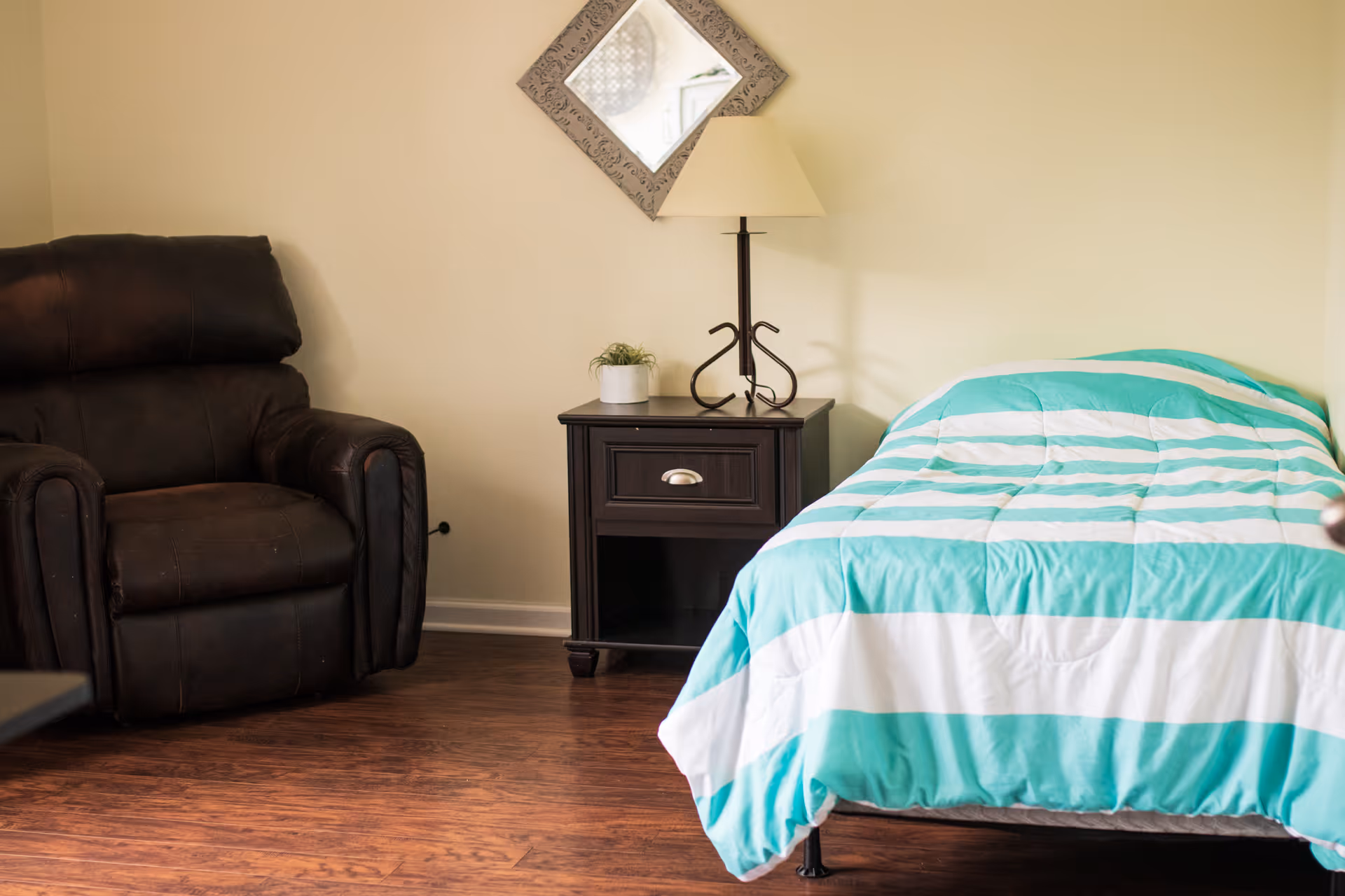 A cozy senior living bedroom featuring a single bed with a turquoise and white striped comforter, a dark brown leather recliner chair, a dark wooden nightstand with a small potted plant and a lamp, and a decorative square mirror hanging on the beige wall.
