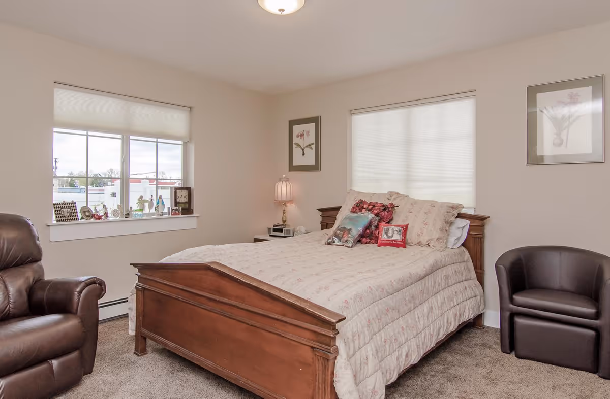 A cozy bedroom with a wooden bed frame and a floral-patterned comforter. There are decorative pillows on the bed, a nightstand with a lamp and clock, two framed floral pictures on the walls, a leather armchair on the left, and a black upholstered chair on the right. Two windows with blinds allow natural light into the room.