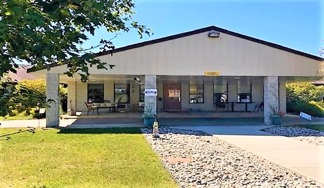 Front exterior view of a single-story building with a covered entrance supported by stone pillars. There are chairs and tables on the porch area, a grassy lawn on the left, and a rock-covered walkway on the right under a clear blue sky.