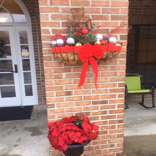 A brick column decorated with a festive holiday basket containing red and silver ornaments, greenery, pine cones, and a large red bow. Below the basket, there is a planter with red poinsettia flowers. To the left, there is a white door with glass panels, and to the right, a green chair is visible on a concrete porch.