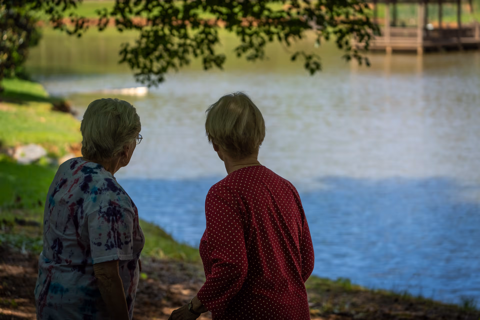 Two elderly women standing near the edge of a lake, looking out over the water with trees and greenery in the background.