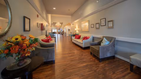 A spacious and well-lit interior hallway with wooden flooring, featuring comfortable seating areas with sofas and armchairs adorned with colorful cushions. A table with a vase of flowers is visible on the left side, and framed pictures hang on the walls. The hallway leads to a dining area with a chandelier in the background.
