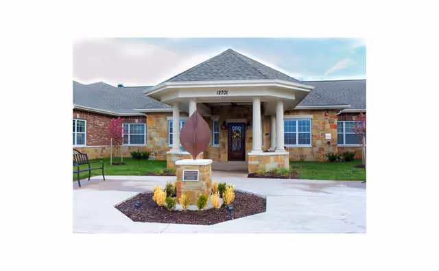 Front exterior view of Silverado Overland Park Memory Care Community building with a covered entrance supported by columns, stone and brick facade, landscaped area with a metal leaf sculpture in front, and a bench on the left side.