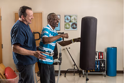 An elderly man in a striped shirt is practicing punching a black punching bag with the assistance of a caregiver in a room equipped with exercise and therapy equipment.