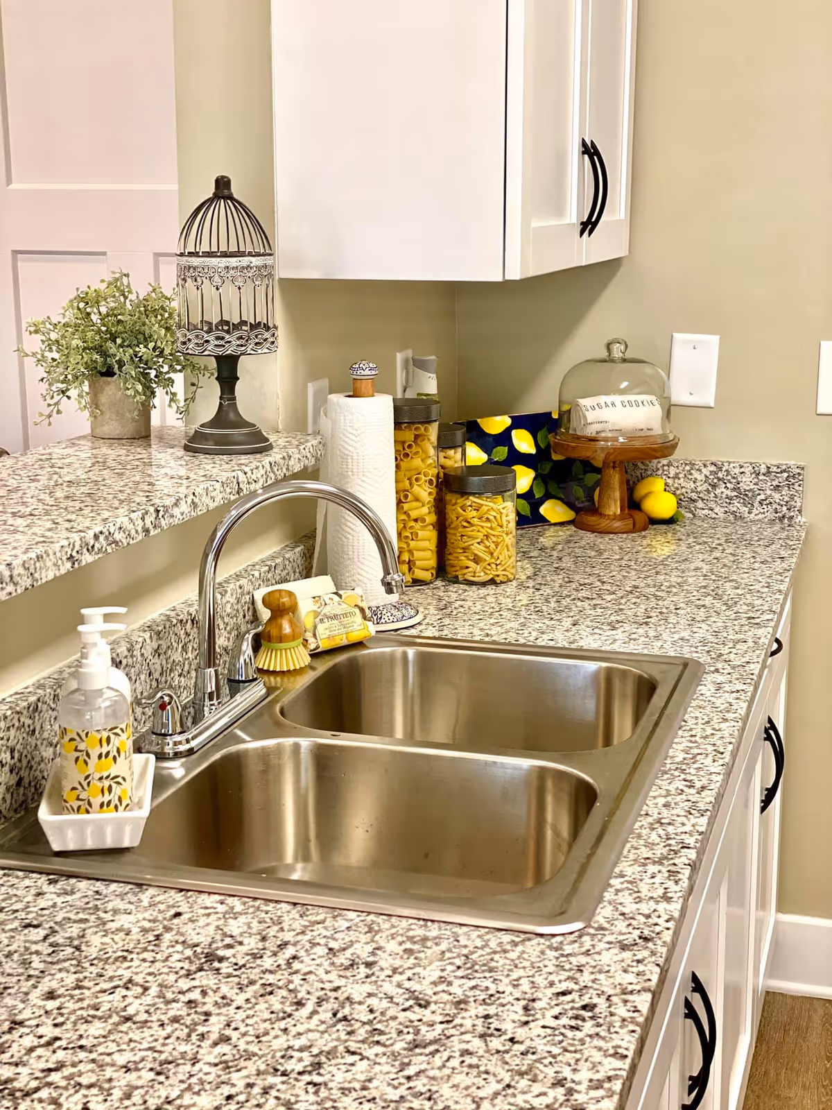 A kitchen countertop with a double stainless steel sink and a chrome faucet. On the counter, there is a soap dispenser with a lemon pattern, a small scrub brush, a bar of soap, a paper towel roll, three jars filled with pasta, a decorative birdcage, a small potted plant, and a glass cake stand with a dome labeled 'Sugar Cookies' containing cookies. The cabinets above and below the counter are white with black handles.