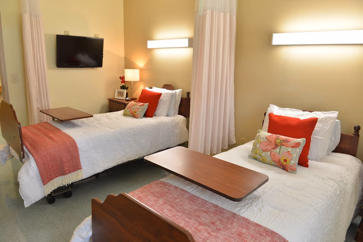 A room with two single beds, each with white bedding, red and floral pillows, and a red throw blanket. Each bed has a wooden overbed table. There is a nightstand between the beds with a lamp, a small plant, and a framed photo. A flat-screen TV is mounted on the wall, and a curtain hangs between the beds.