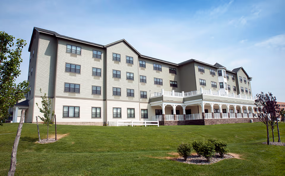 Exterior view of a multi-story senior living facility building with beige siding and multiple windows. The building has a large porch with white railings and is surrounded by a well-maintained green lawn with small trees and shrubs under a partly cloudy sky.