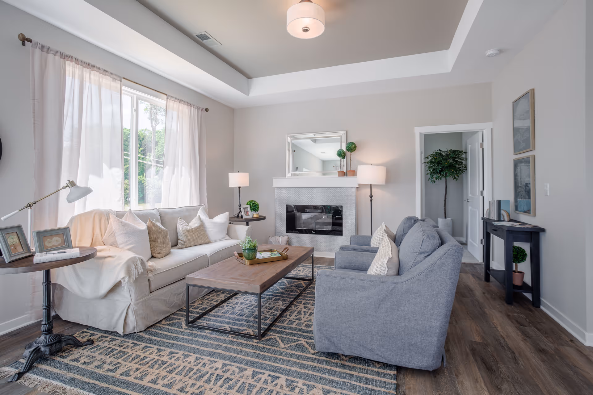 A bright and cozy living room with a large window covered by sheer curtains letting in natural light. The room features a beige sofa with multiple cushions, two gray armchairs, a wooden coffee table with a small plant and tray, a patterned area rug, and a fireplace with a mirror and decorative plants above it. There are two floor lamps on either side of the fireplace, a small round side table with framed photos and a lamp, and a black console table with decorative items and a potted plant. The walls are painted light gray and the floor is wood.
