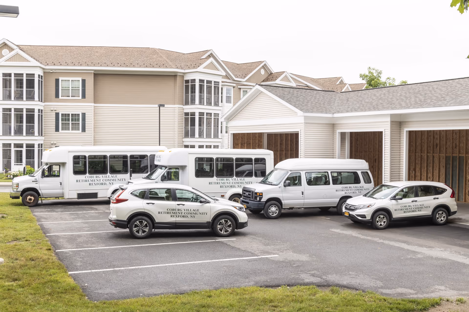 Parking area outside a senior living facility with two white shuttle buses and two white SUVs, all labeled 'Coburg Village Retirement Community Rexford, NY'. The background shows a multi-story residential building with beige siding and multiple windows.