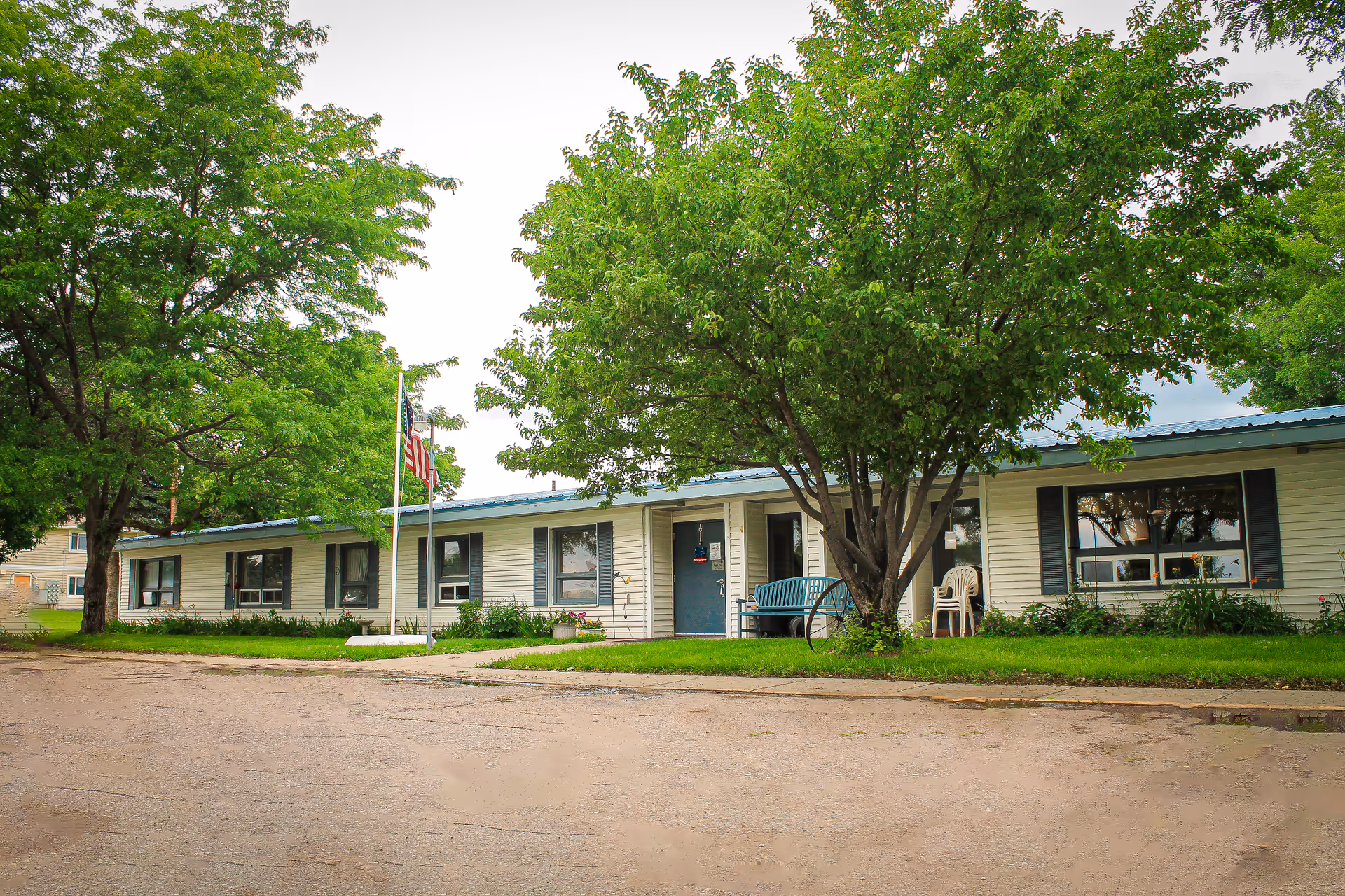 Single-story building with white siding and blue trim surrounded by green trees and grass. There is a blue bench and a white plastic chair near the entrance, and an American flag on a flagpole in front of the building.