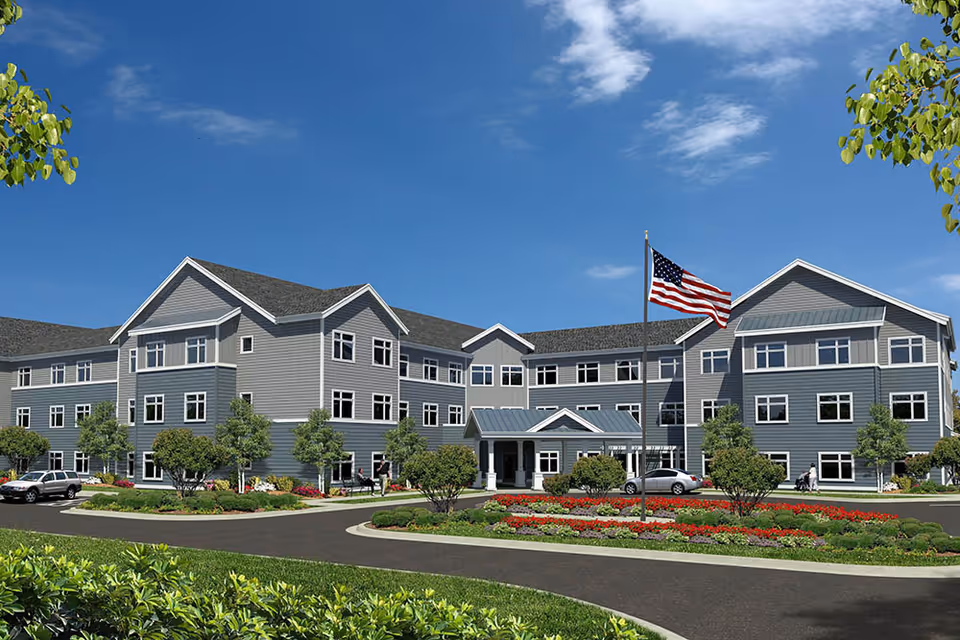 Exterior view of a large, modern three-story senior living facility with gray siding and white trim. The building is surrounded by landscaped gardens with green shrubs and colorful flowers. An American flag flies on a flagpole in front of the entrance, and a few cars are parked nearby under a clear blue sky.