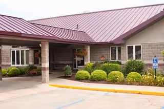 Entrance area of a care facility with a covered drop-off zone supported by stone pillars, a maroon metal roof, beige brick walls, green shrubs, and a handicapped parking sign near the sidewalk.